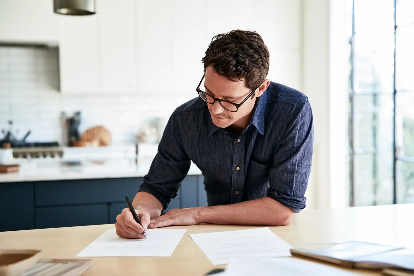 Man in his 30s signing a single document at a bright kitchen table — applying for a makwa loan at home